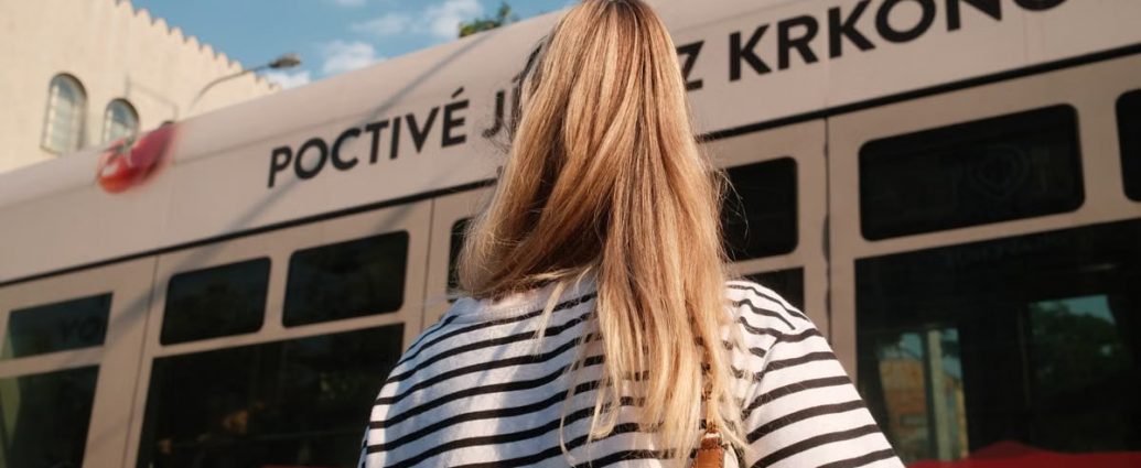 A woman with blonde hair in a striped shirt stands near a city tram on a sunny day.