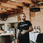 Man in rustic winery setting with bottles and wooden barrels, embracing a casual vibe.