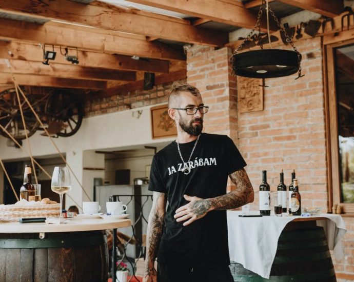Man in rustic winery setting with bottles and wooden barrels, embracing a casual vibe.